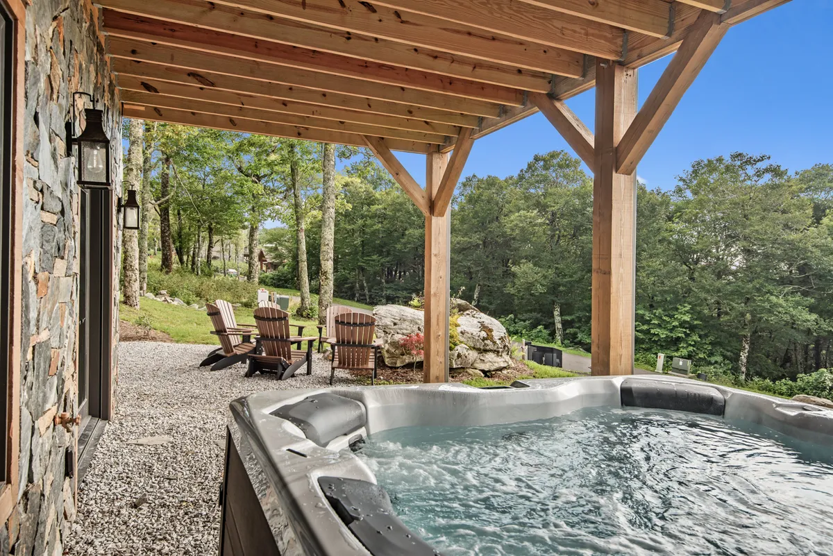A serene outdoor area featuring a BullFrog hot tub surrounded by wooden chairs and a stone wall, nestled among trees. The setting offers sunset views from the hot tub, with a cozy ambiance perfect for soaking and socializing.