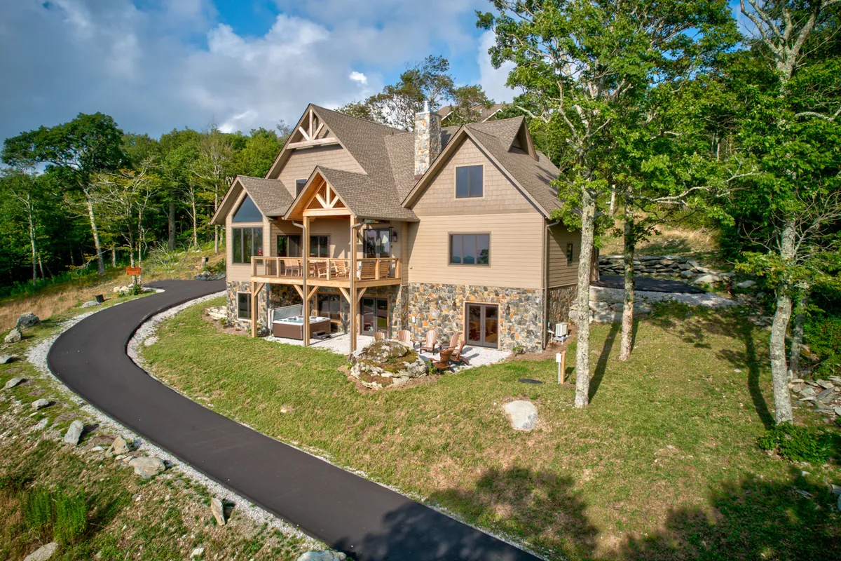 The image showcases the grand stone facade of the home, framed by lush greenery and an expansive wraparound porch. The scene is tranquil, bathed in the soft glow of sunset, inviting guests to imagine sipping coffee or relaxing with loved ones while taking in panoramic mountain views.