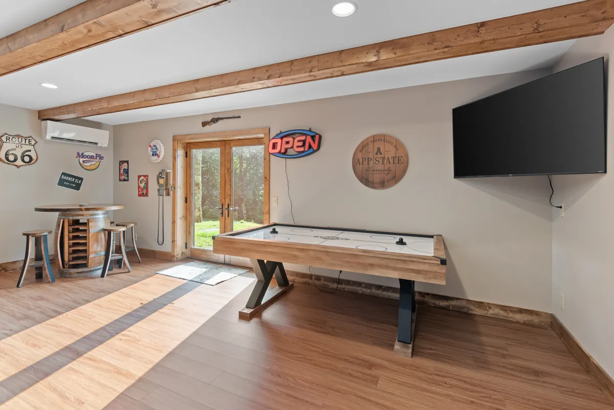 The image displays an air hockey table and a barrel-style table set beneath wooden beams, with an open door letting in natural light and a neon 'OPEN' sign adding a playful vibe. The layout suggests a space ideal for both action-packed games and relaxed gatherings.