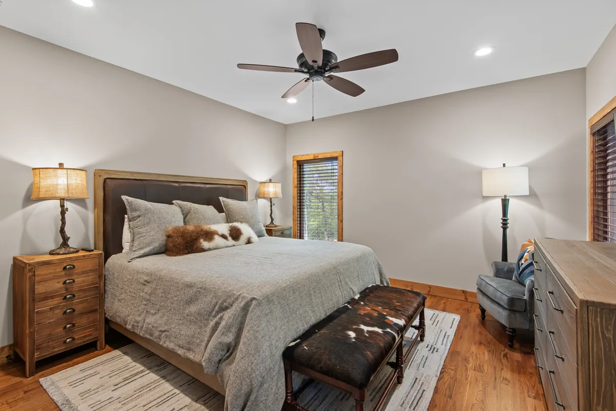 A tranquil bedroom featuring a bed with gray bedding, wooden bedside tables, and a decorative bench at the foot. The window with blinds allows in soft, natural light, creating a serene and restful atmosphere.