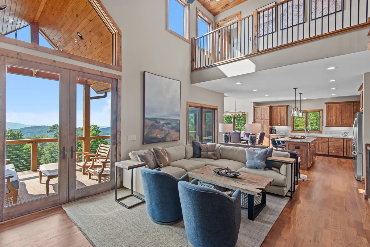 This image features a spacious, welcoming open-concept area with a large window framing expansive mountain views. The space includes a comfortable seating area, a wooden dining table, and a modern kitchen with appliances visible. Natural light fills the room, and the seamless flow between living, dining, and kitchen spaces is evident.