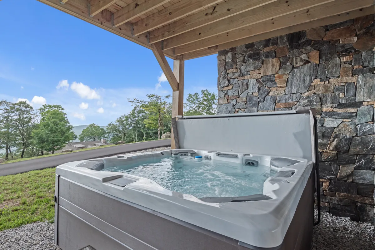 This exterior scene features a tranquil hot tub set against a stone wall and wooden structure, with a beautiful landscape view in the background. The mood is peaceful, perfect for relaxing, entertaining guests, or simply soaking while enjoying nature or a sunset.
