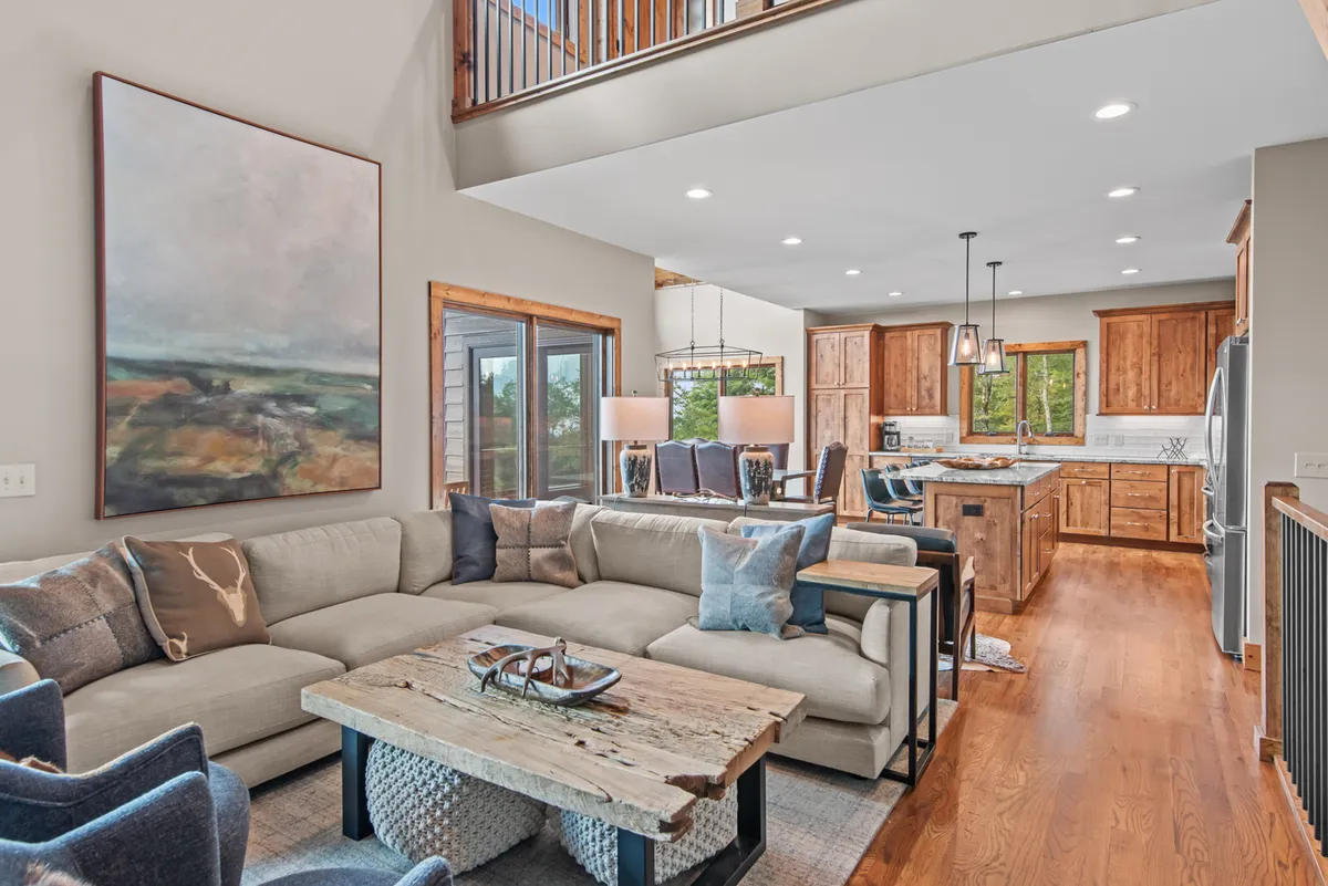 An inviting living area with a large sectional sofa, rustic coffee table, kitchen island, and artwork on the wall. The open concept design is airy and welcoming, ideal for relaxing or entertaining.