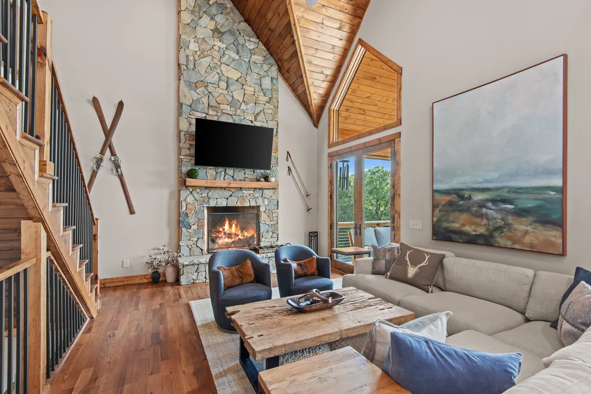 A warm, inviting living room scene anchored by a substantial stone fireplace. Large windows let in natural light and offer glimpses of the mountain. Cozy armchairs, a coffee table, and decorative touches create a welcoming atmosphere for groups.